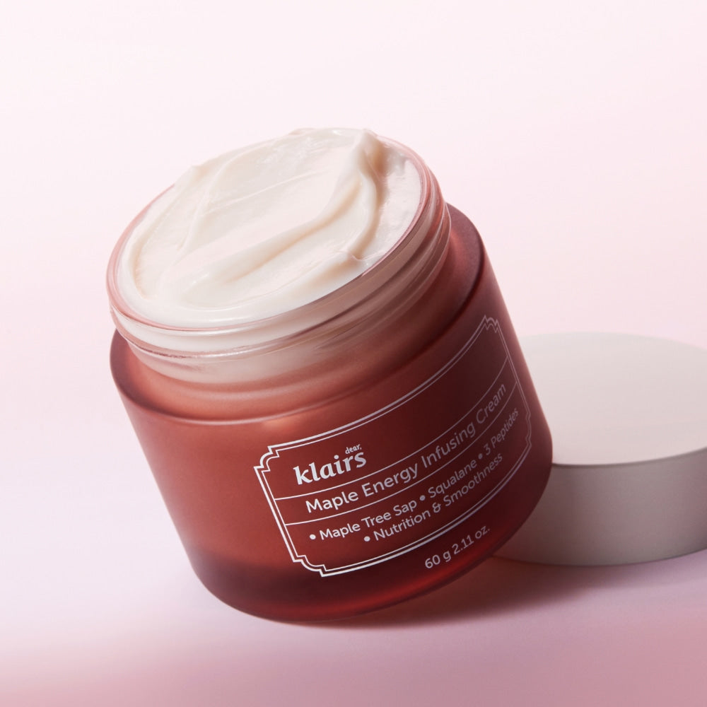 Open jar of KLAIRS Maple Energy Infusing Cream 60g with white cream and labeled brown container on pink background.