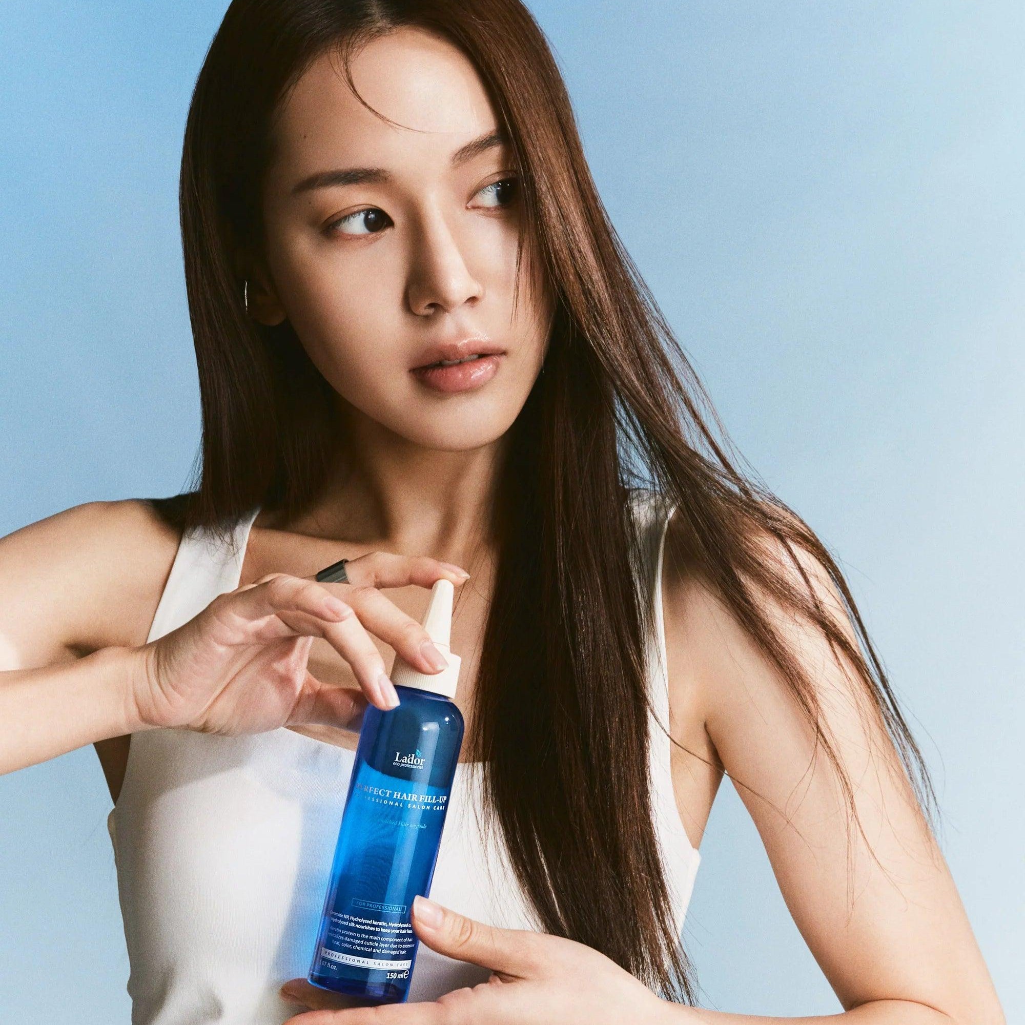 Woman holding LADOR Perfect Hair Fill-Up 150ml bottle against blue background