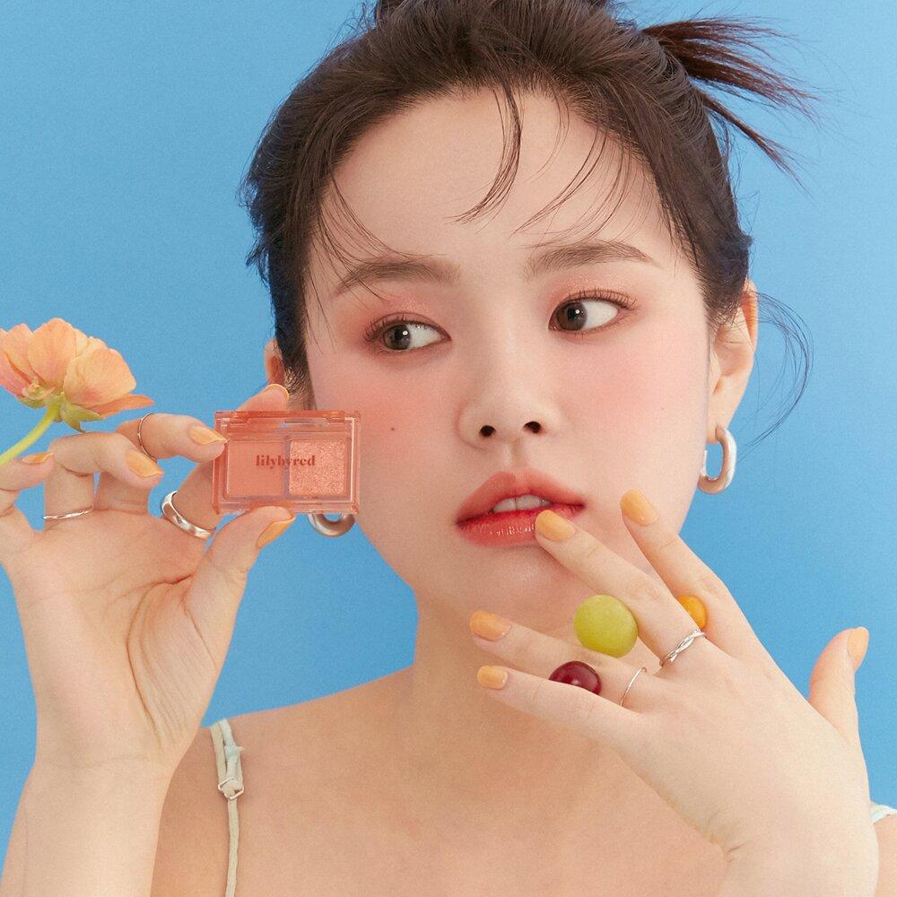 Woman showcasing LILYBYRED Little Bitty Moment Shadow against a blue background with orange flower and vibrant nail polish.