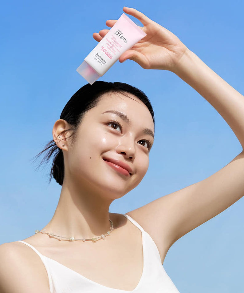 Smiling person holding MAKE P:REM Soothing Pink Tone-Up Sunscreen SPF 50+ PA++++ 50ml against a clear blue sky.