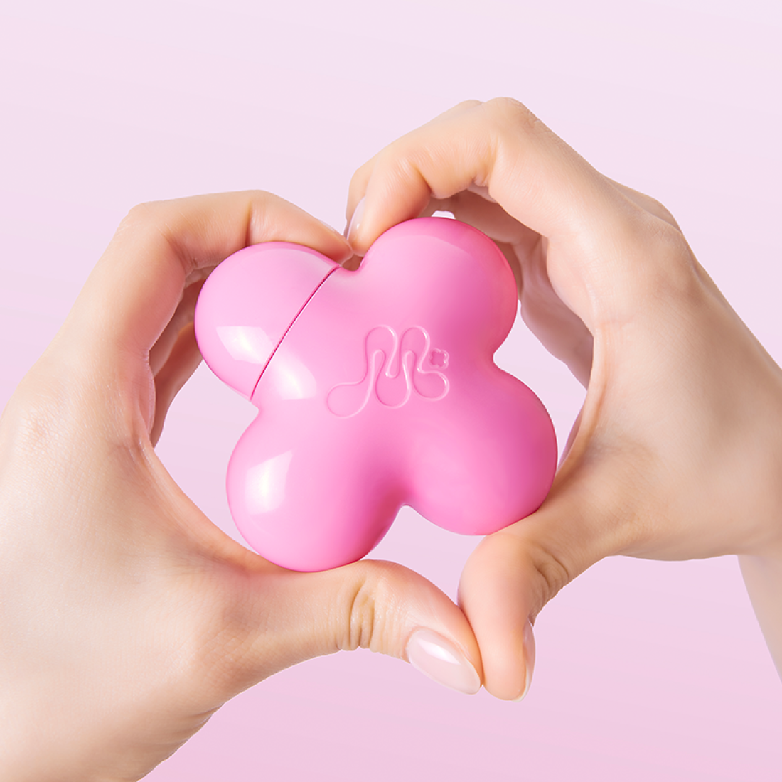 Hands holding pink clover-shaped beauty product against a pink background.