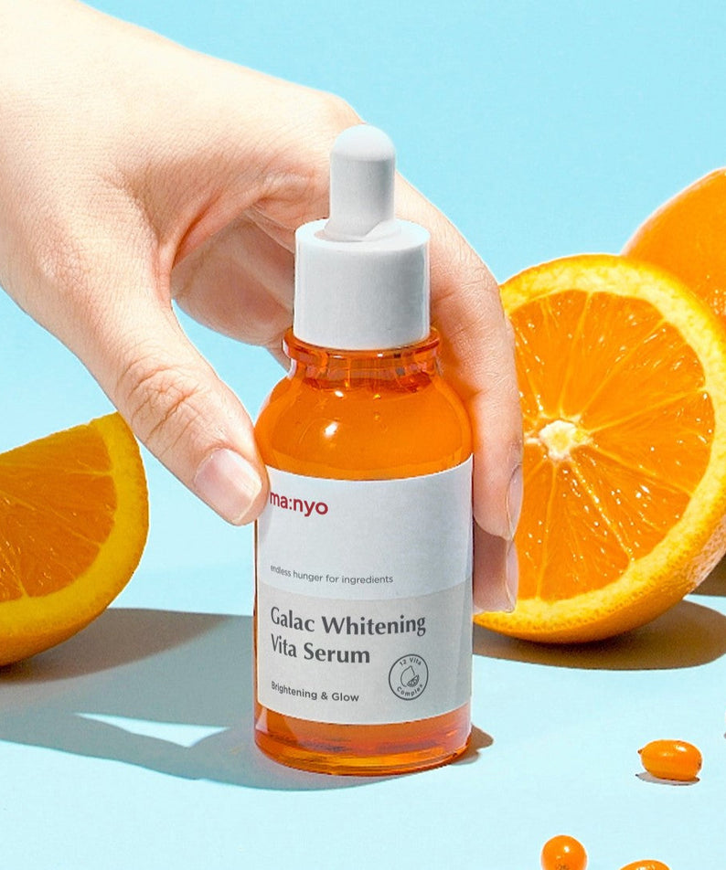 Hand holding MA:NYO Galac Whitening Vita Serum 50ml with sliced oranges in the background, showcasing the product's brightening effect.