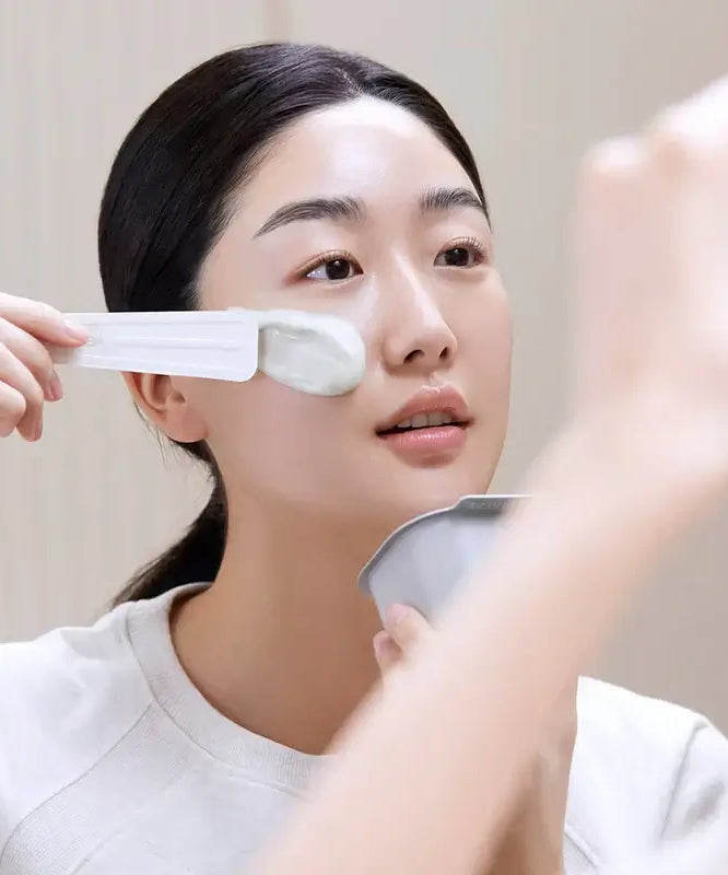 Woman applying MEDIHEAL Derma Modeling Pack [Hyaluronic Acid Moisturizing] to her face with a spatula