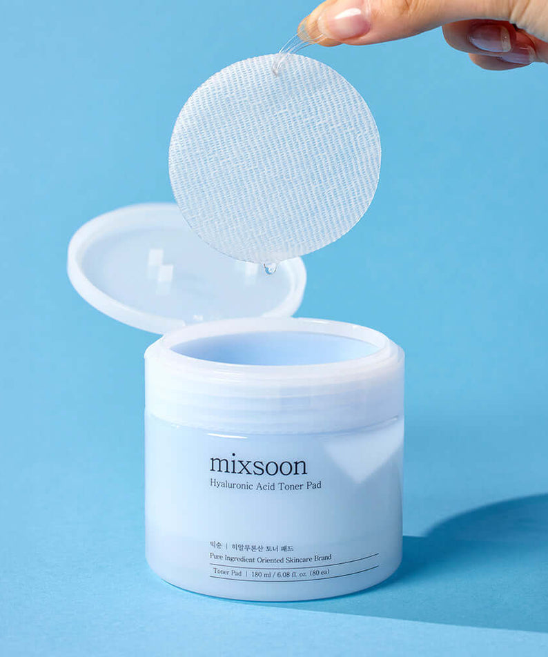 Hand holding a MIXSOON Hyaluronic Acid Toner Pad above an open jar on a blue background.