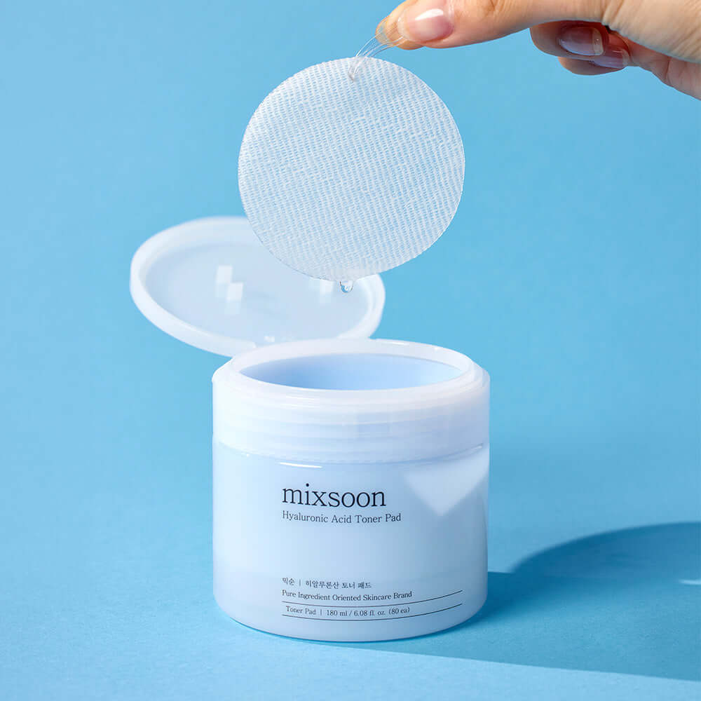 Hand holding a MIXSOON Hyaluronic Acid Toner Pad above an open jar on a blue background.