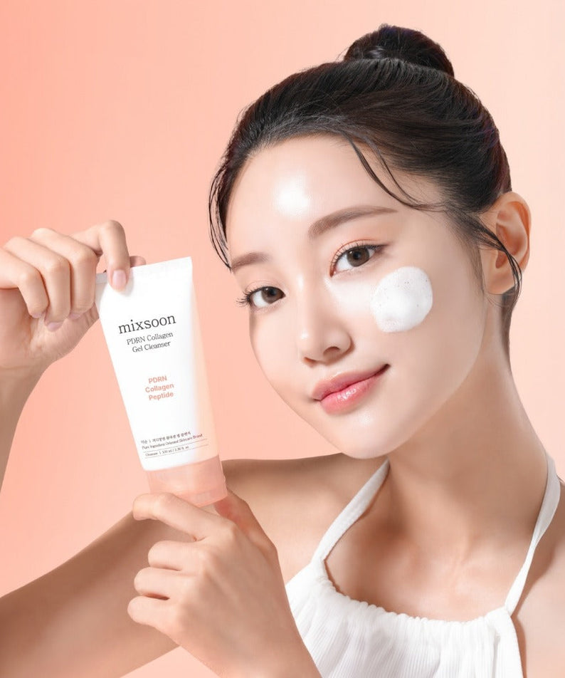 Person holding MIXSOON PDRN Collagen Gel Cleanser 100ml with a small amount applied to their cheek, smiling against a peach background.