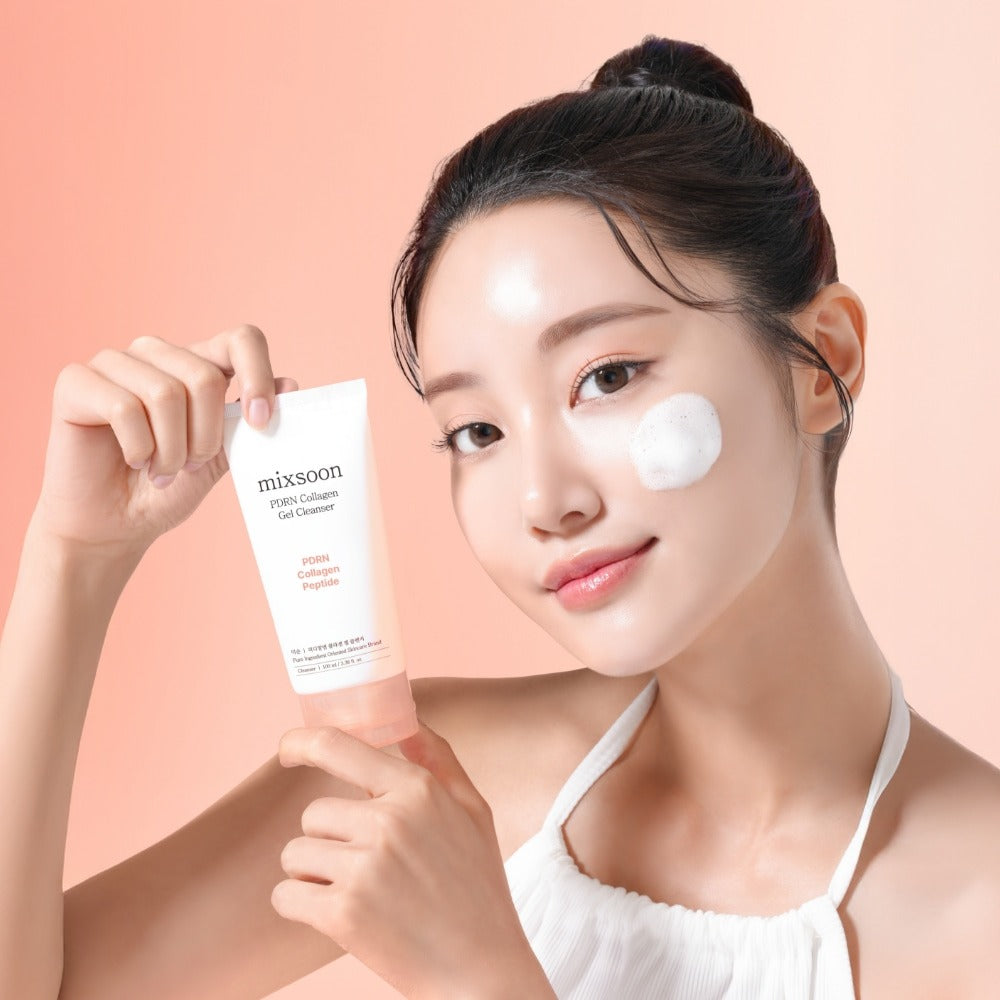 Person holding MIXSOON PDRN Collagen Gel Cleanser 100ml with a small amount applied to their cheek, smiling against a peach background.