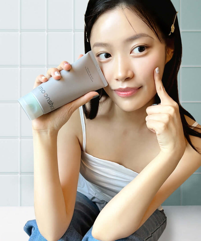 Woman holding MOONSHOT Micro Black Cleansing Foam tube against her cheek in a bathroom.
