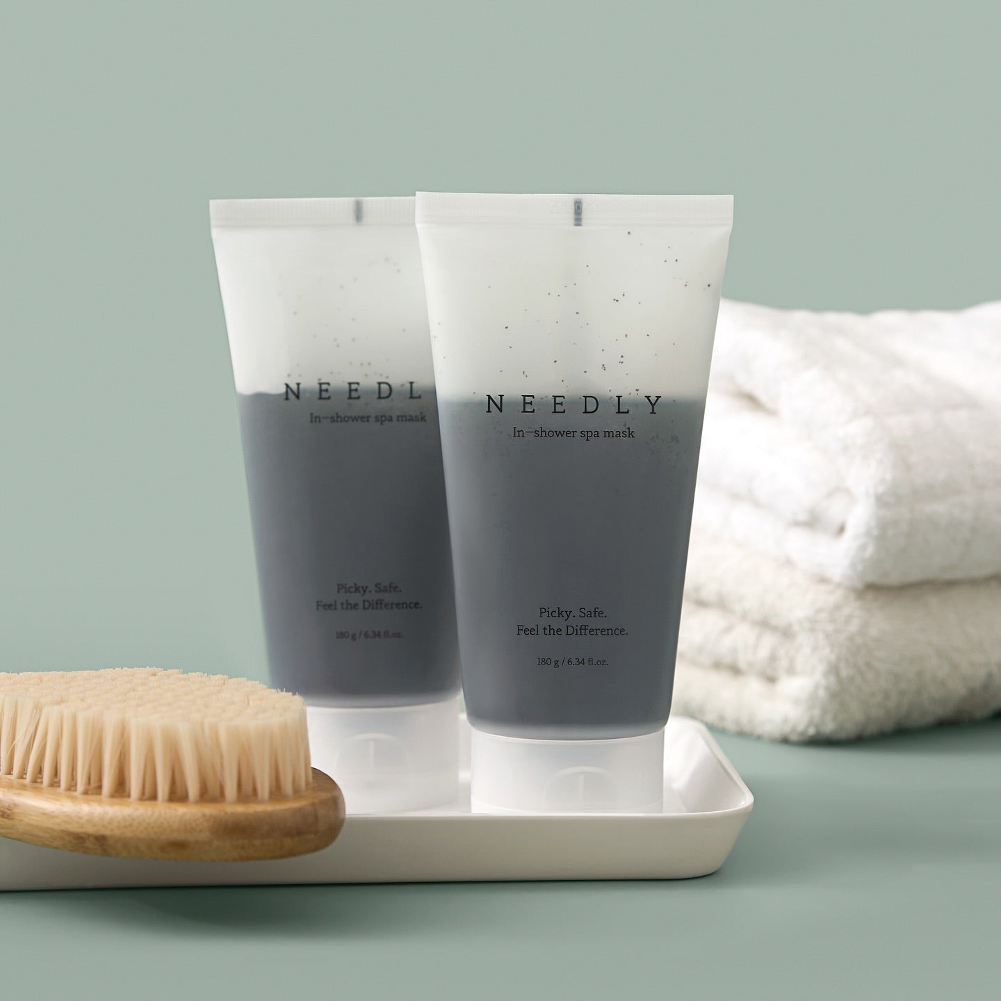 Two tubes of NEEDLY Ishower Spa Mask 180g next to a dry brush and towels, highlighting spa-like skincare experience.