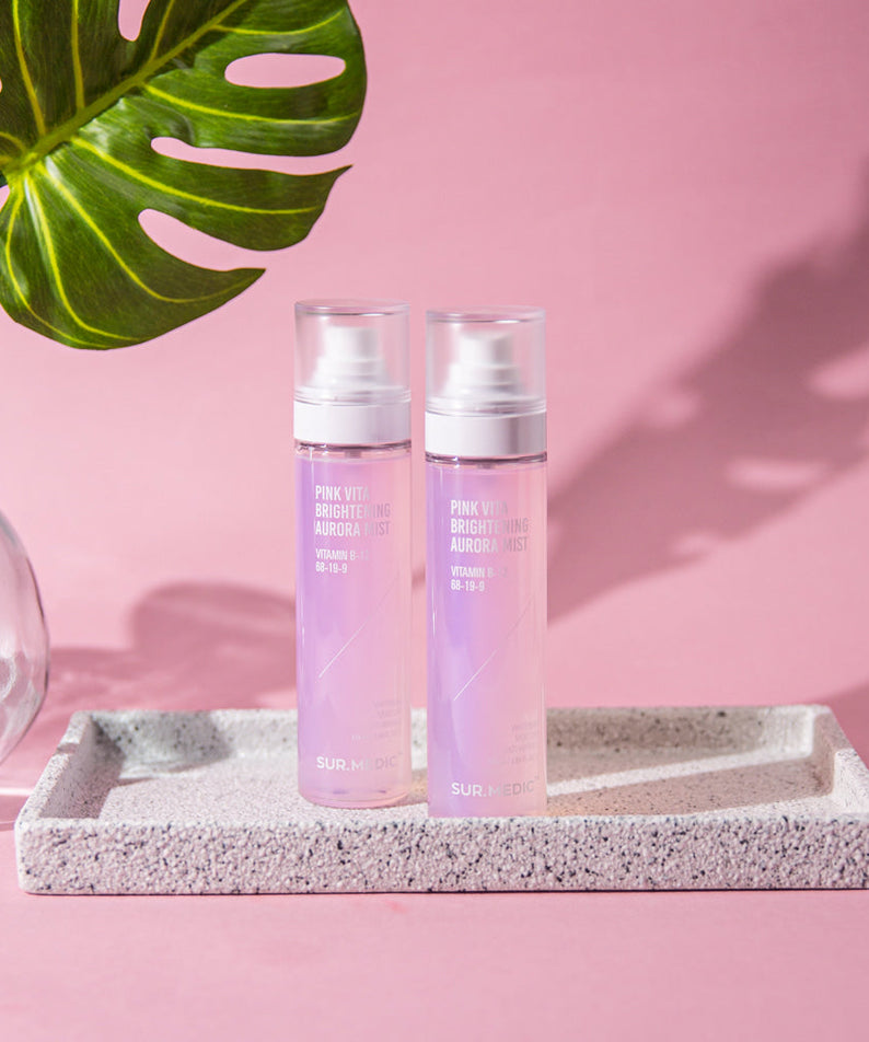 Two bottles of NEOGEN Pink Vita Aurora Mist 115ml on a tray with a palm leaf and pink background, showcasing product design.