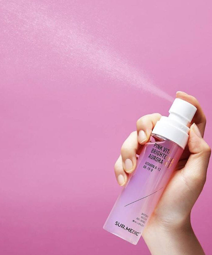 Hand holding NEOGEN Pink Vita Aurora Mist 115ml against pink background, spraying fine mist