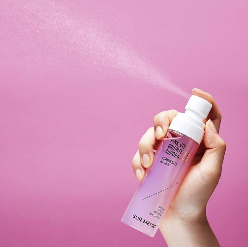 Hand holding NEOGEN Pink Vita Aurora Mist 115ml against pink background, spraying fine mist