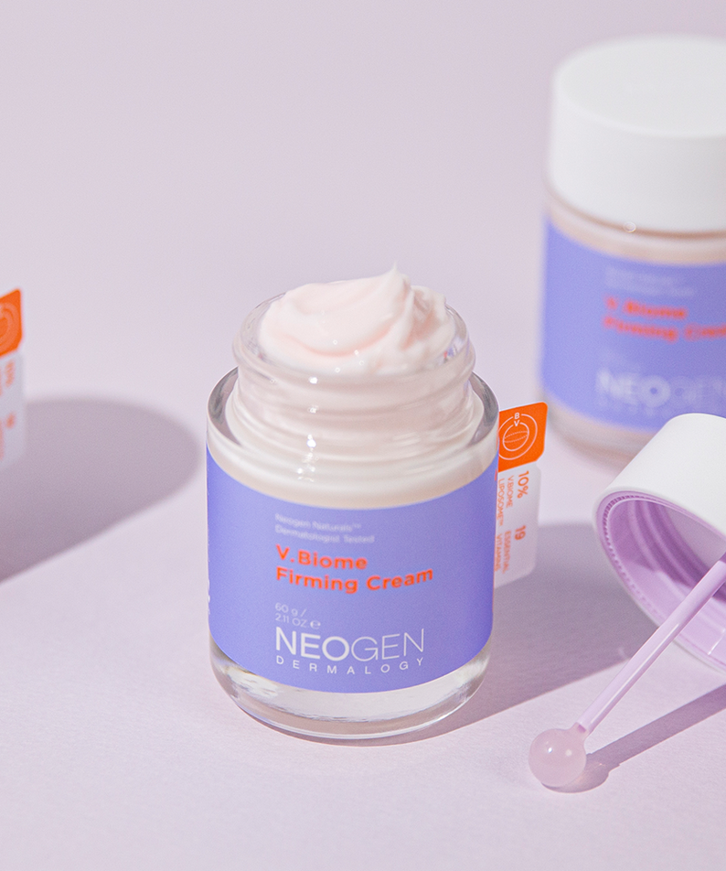 Open jar of NEOGEN V Biome Firming Cream 60g on a light pink background with lid and spatula.