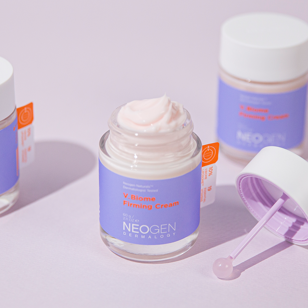Open jar of NEOGEN V Biome Firming Cream 60g on a light pink background with lid and spatula.