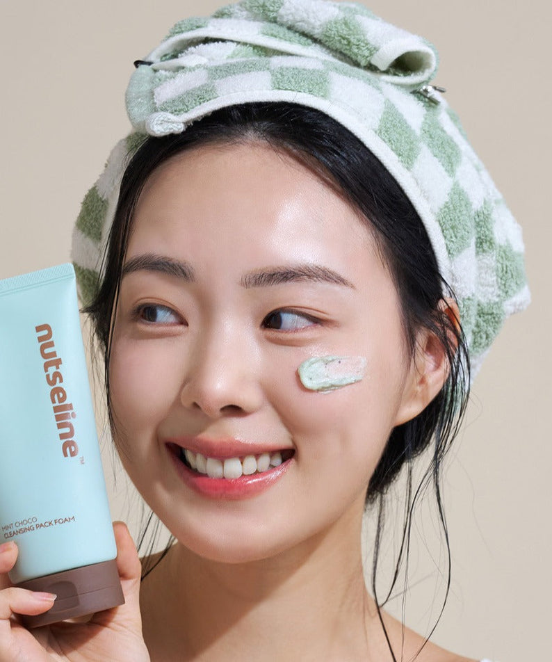Smiling woman applying NUTSELINE Mint Chocolate Cleansing Pack Foam 130g with towel on head.