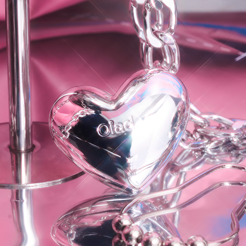 Silver heart-shaped pendant with 'oiad' engraving on a chain, set against a pink reflective background.