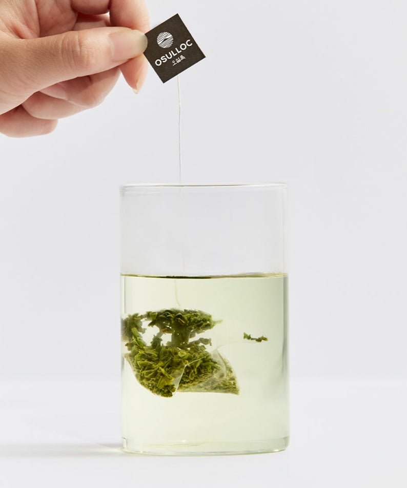 Brewing OSULLOC Sejack Green Tea 20 P tea bag in a glass with fresh green leaves visible.