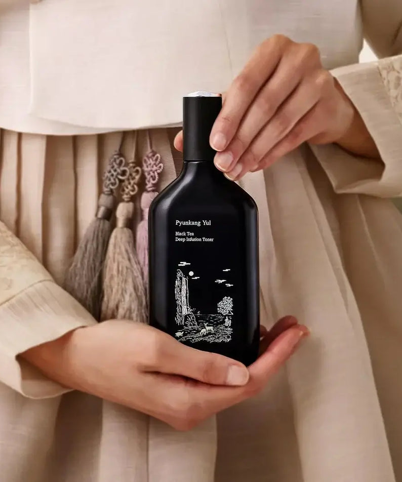 Hands holding PYUNKANG YUL Black Tea Deep Infushion Toner 130ml bottle with elegant design packaging.