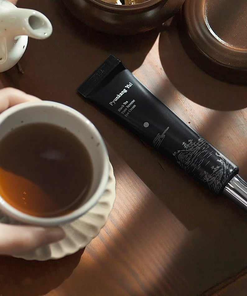 PYUNKANG YUL Black Tea Time Reverse Eye Cream 25ml placed next to a cup of tea on a wooden surface