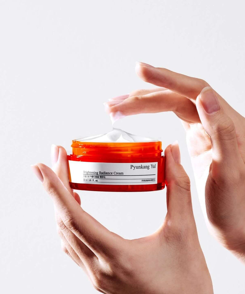 Hands holding PYUNKANG YUL Brightening Radiance Cream 50ml jar, showcasing skincare product texture and packaging.