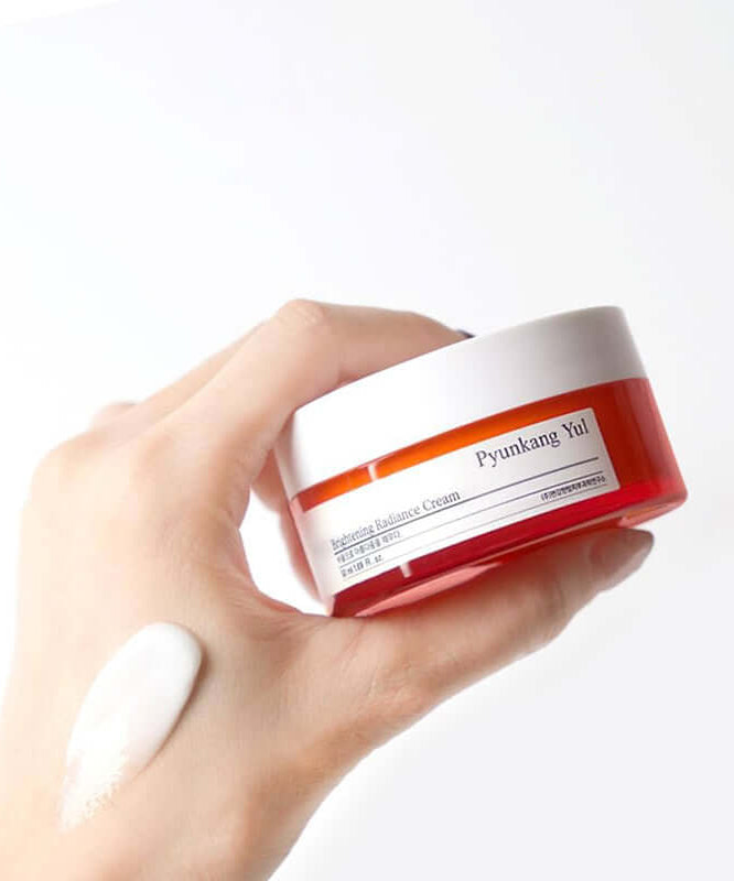 Hand holding a jar of PYUNKANG YUL Brightening Radiance Cream 50ml with cream applied on the skin, showcasing product texture.