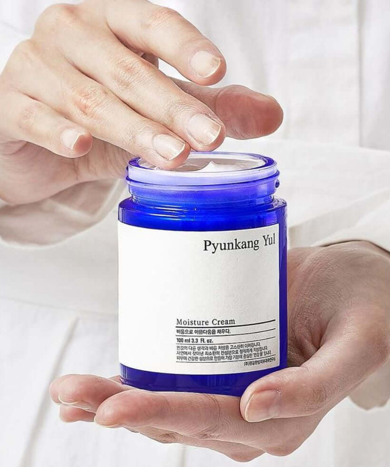 Hands holding an open jar of PYUNKANG YUL Moisture Cream 100ml with a blue container.