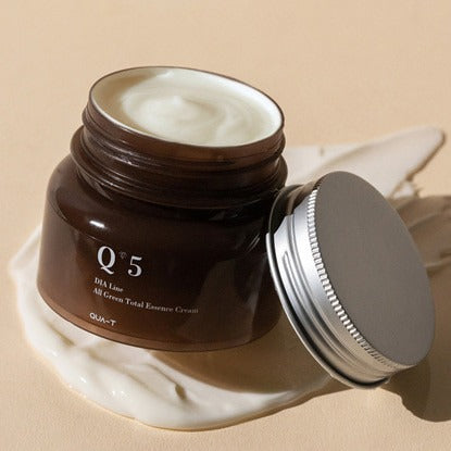 Open jar of QUA-T Dia All Green Total Essence Cream 100ml on a beige background.