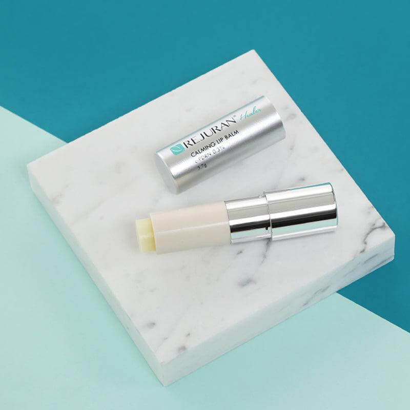 REJURAN Healer Calming Lip Balm 3.7g on marble surface with silver cap removed, showcasing minimalistic design and soothing product features