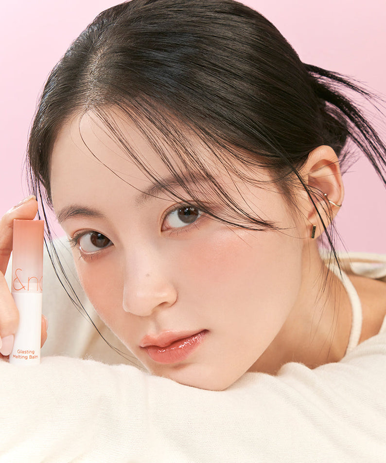 Woman holding ROMAND Glasting Melting Balm (#01 to #15) with smooth complexion, showcasing product's moisturizing effect on lips.