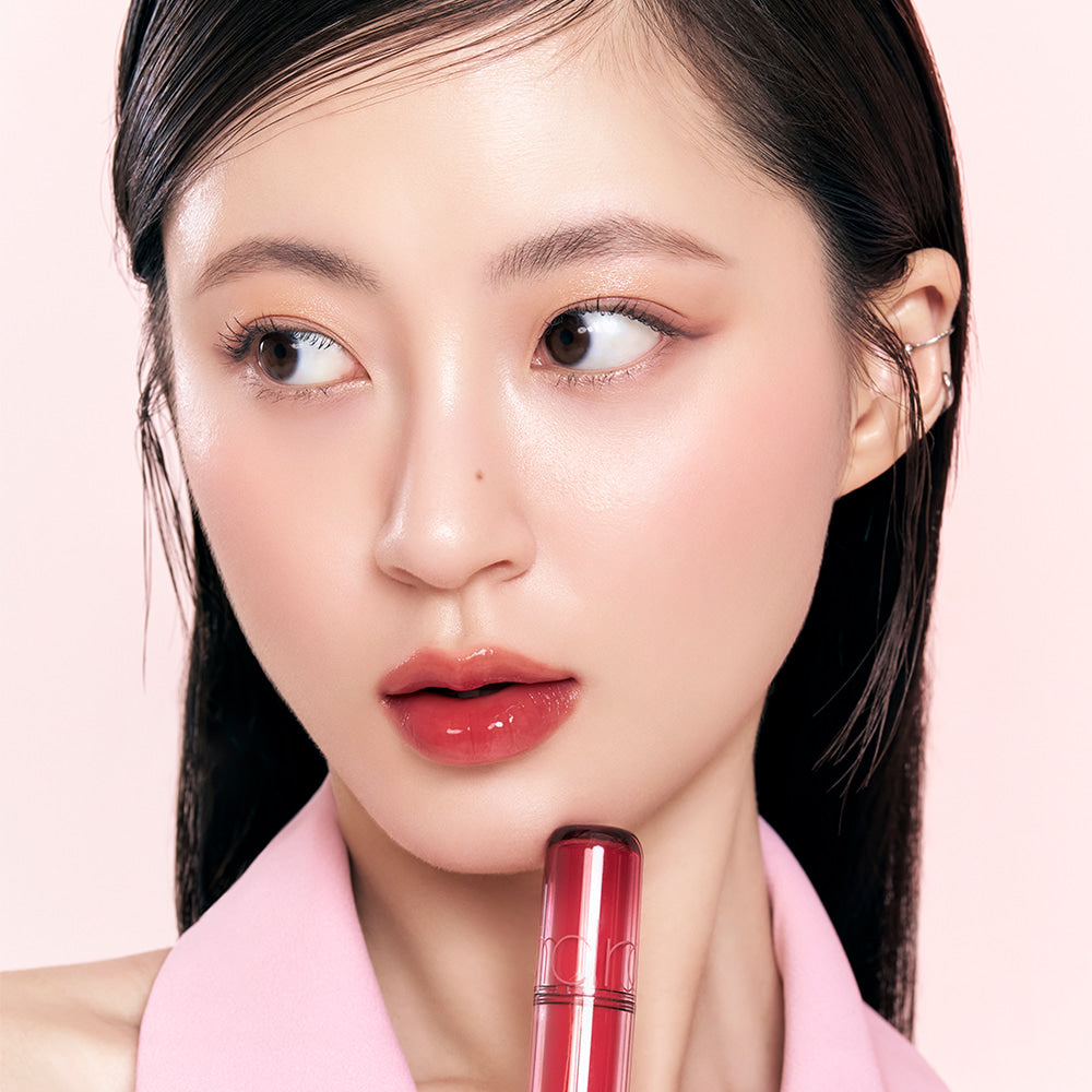 Model applying vibrant ROMAND The Juicy Lasting Tint to lips, showcasing glossy finish and intense color on pale pink background.