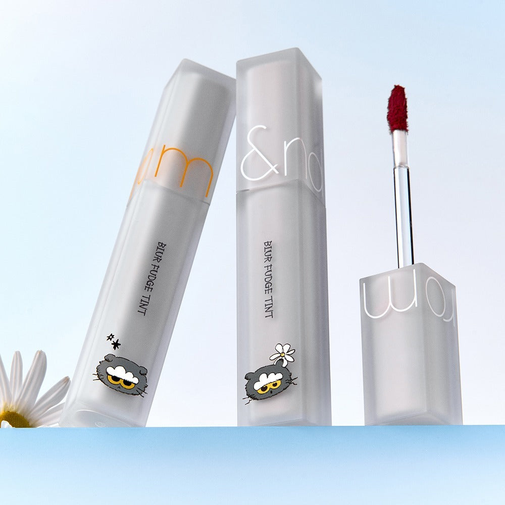 ROMAND x Joanne Friends Blur Fudge Tint makeup product with decorative packaging and lip tint applicator on a light background.
