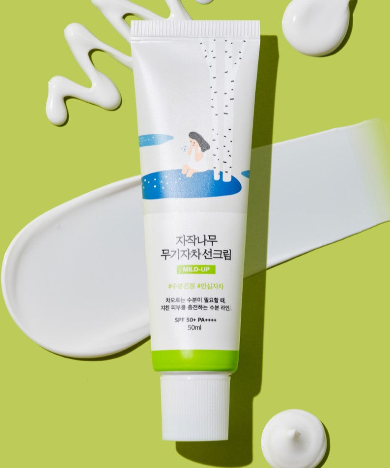 ROUND LAB Birch Juice Moisturizing Mild-Up Sunscreen 50ml on a green background with product swatches