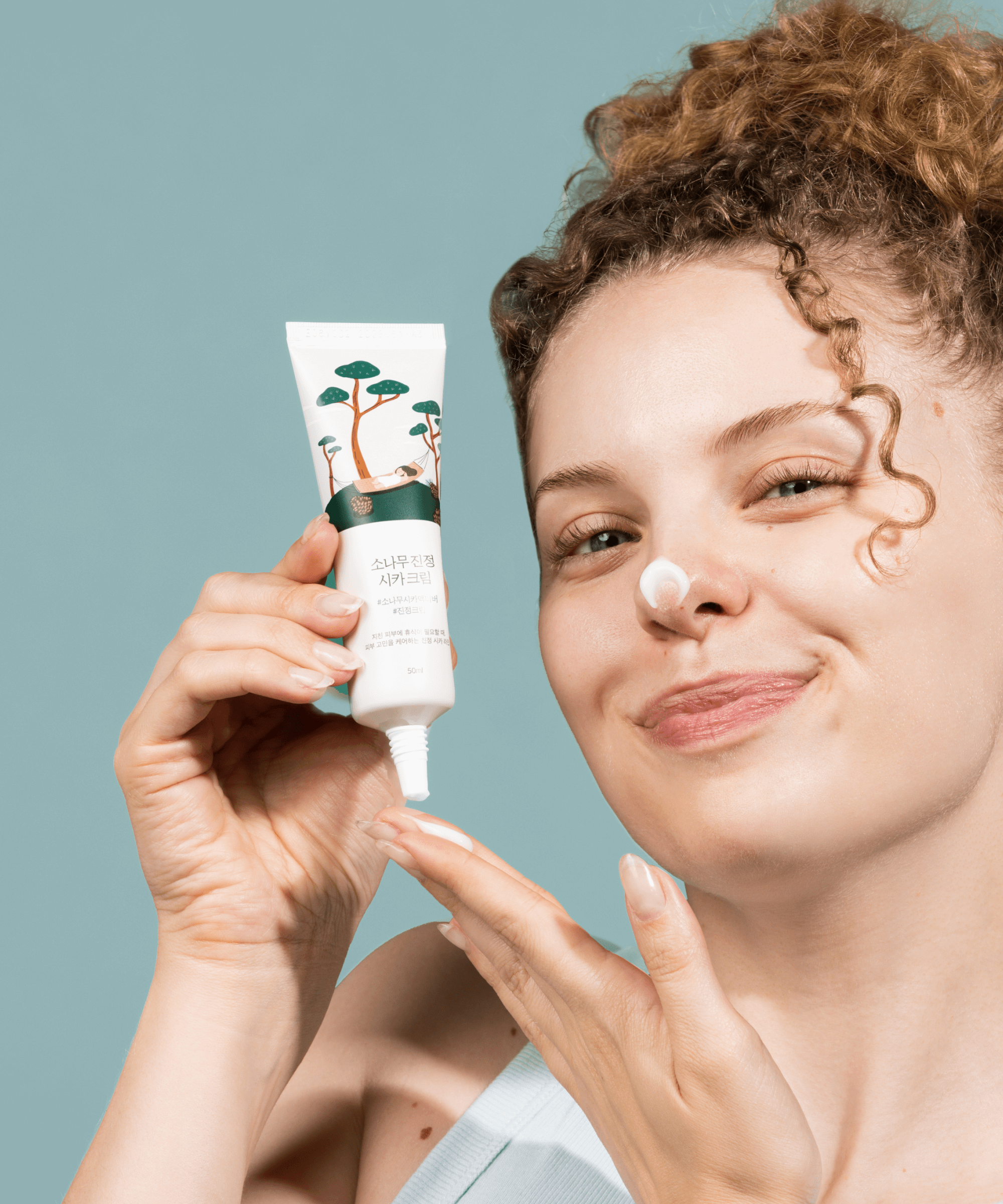 Woman applying ROUND LAB Pine Calming Cica Cream 50ml to her face with a smile, showcasing the skincare product's calming effects.
