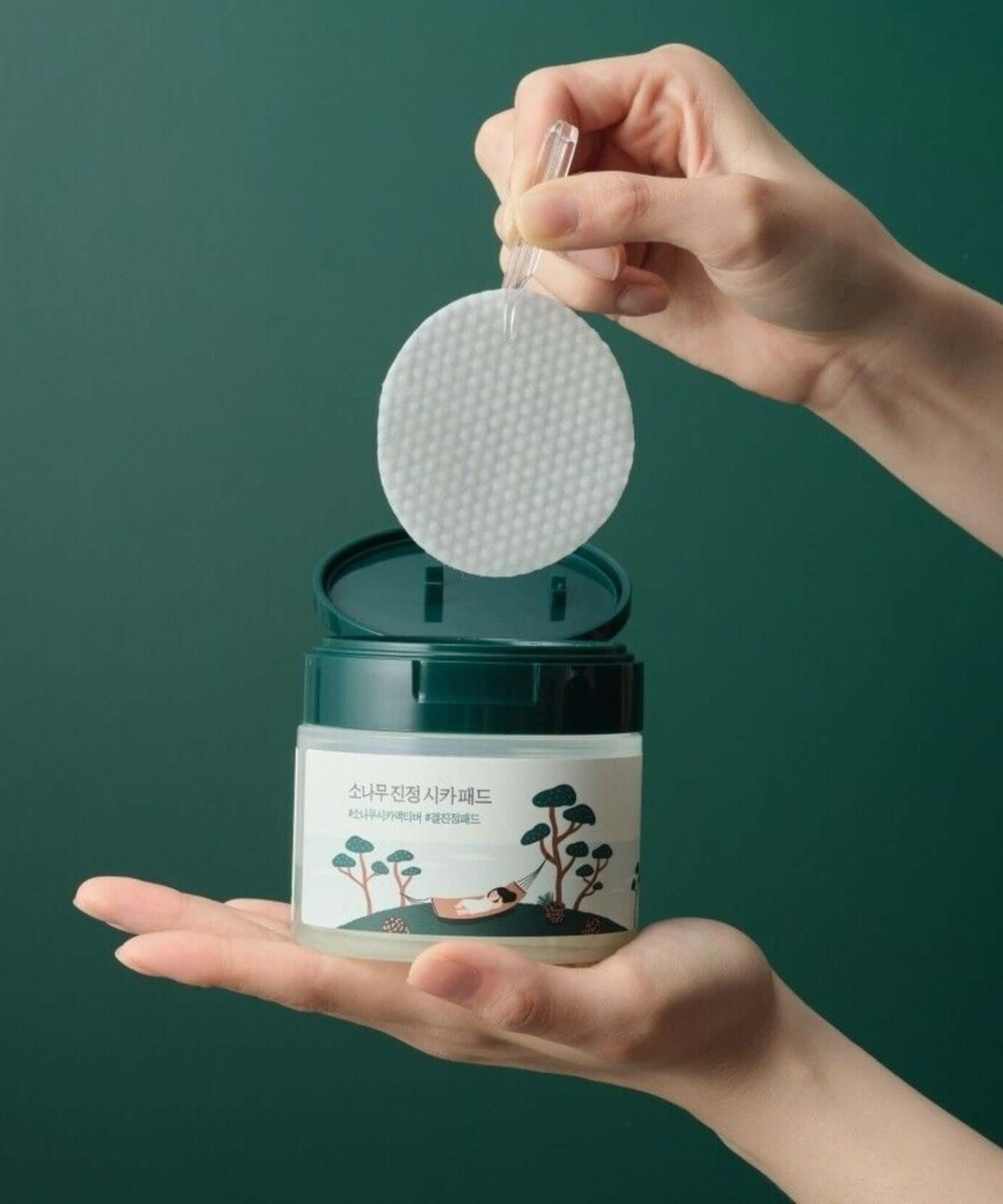 Hands holding ROUND LAB Pine Calming Cica Pads 50 sheets with open jar, showcasing textured pad against green background