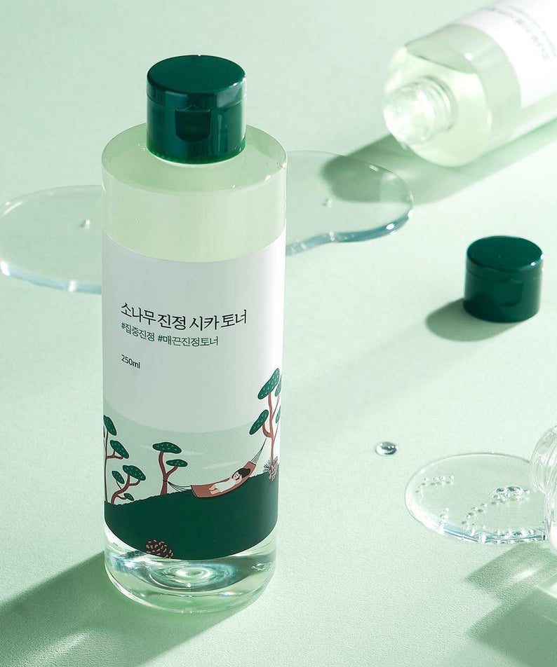 ROUND LAB Pine Calming Cica Toner 250ml bottle on a light green background with scattered toner and open caps. Skincare product image.