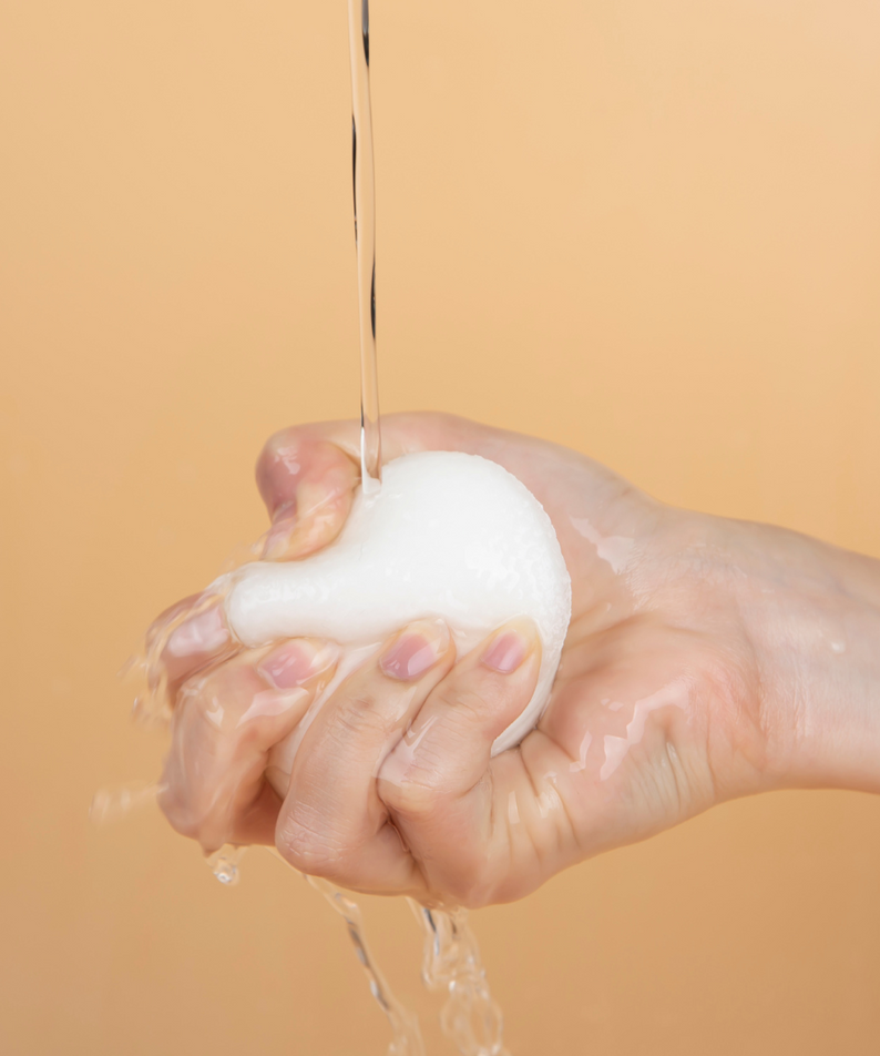 Hand squeezing a foam ball with water dripping against a peach background, illustrating cleansing product application.