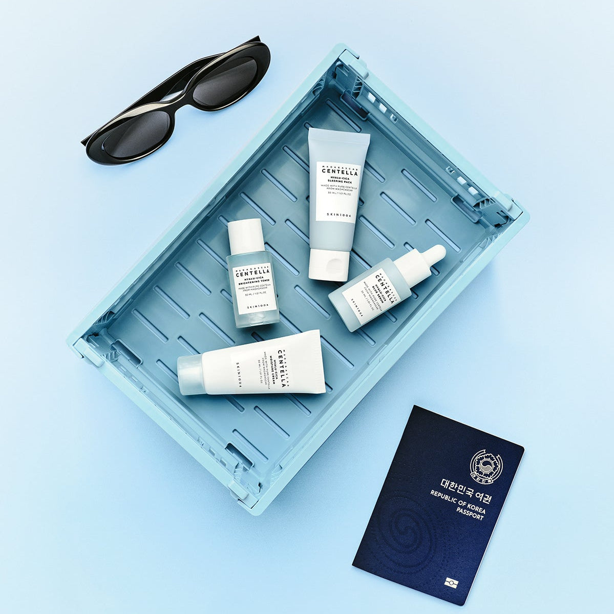 SKIN1004 Madagascar Centella Hyaluronic Acid Travel Kit with skincare products in a blue tray, alongside sunglasses and a passport.