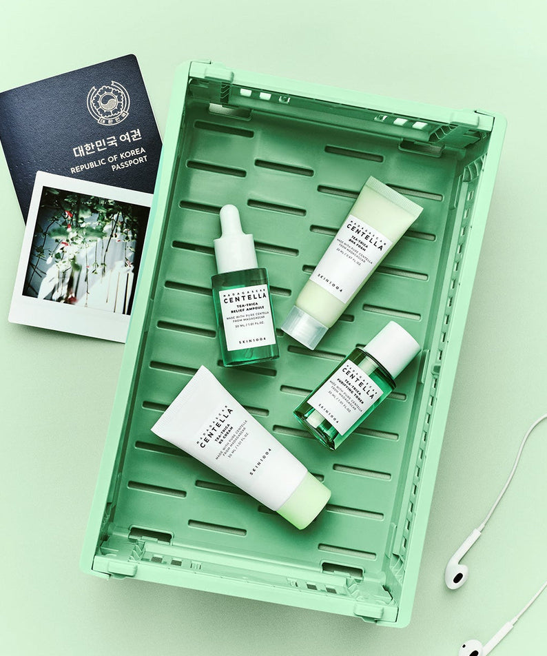 SKIN1004 Madagascar Centella Teatrica Travel Kit displayed in a tray with travel essentials including a passport and headphones.