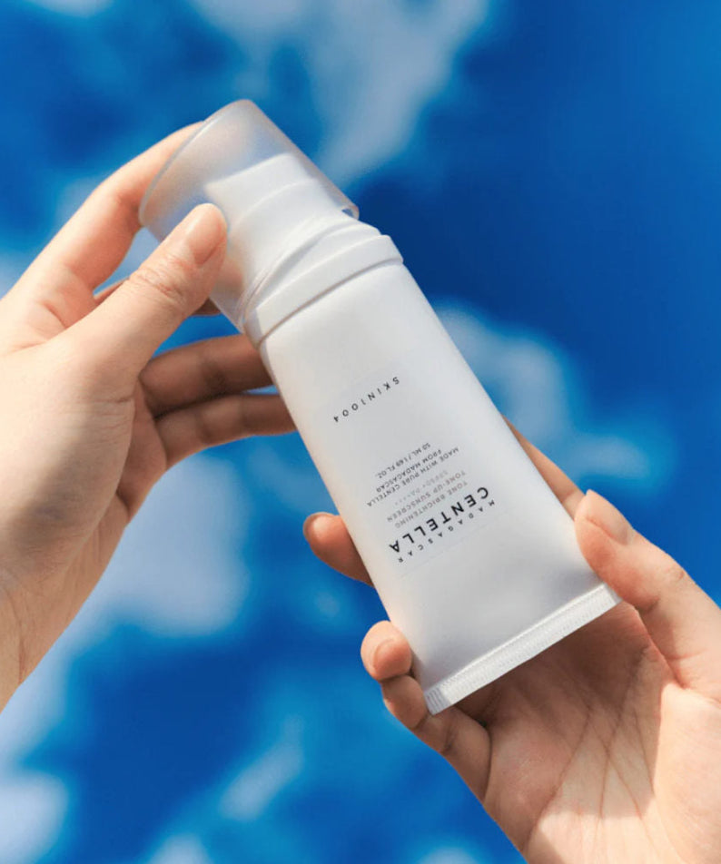 Hands holding SKIN1004 Madagascar Centella Tone Brightening Tone-Up Sunscreen 50ml against a blue sky background