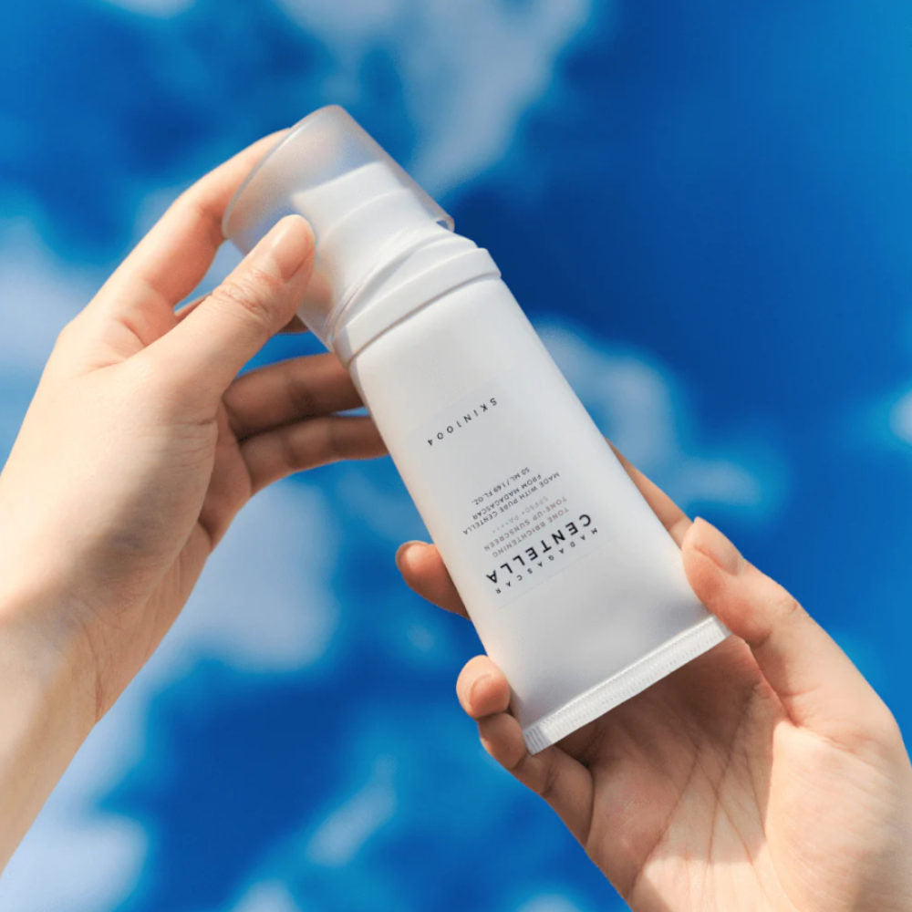 Hands holding SKIN1004 Madagascar Centella Tone Brightening Tone-Up Sunscreen 50ml against a blue sky background