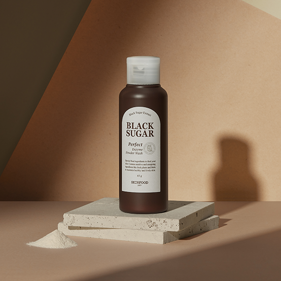 SKINFOOD Black Sugar Perfect Enzyme Powder Wash 65g bottle on a beige background with a shadow and powder spill.
