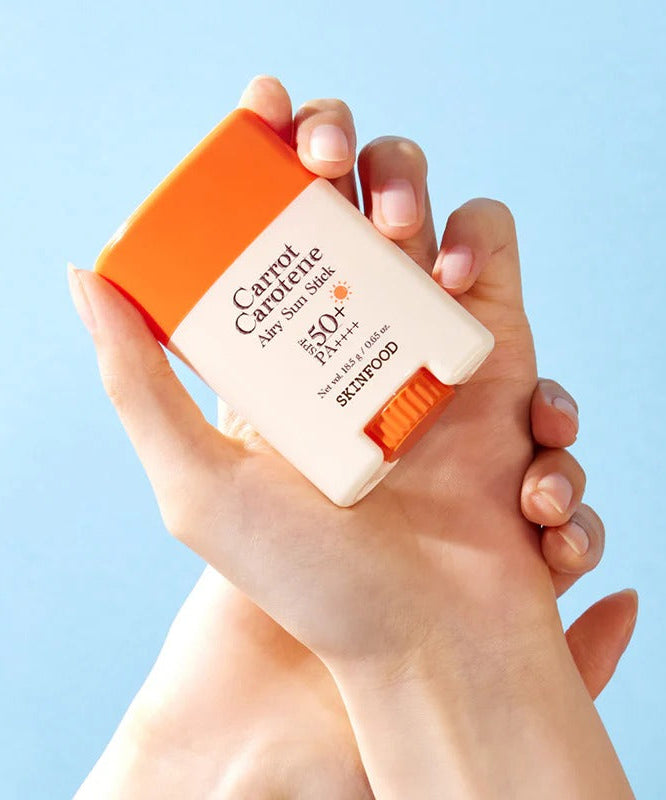 Person holding SKINFOOD Carrot Airy Soft Sun Stick 18.5g with SPF 50 against a blue background.