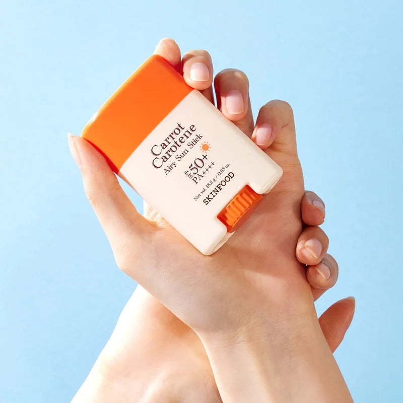 Person holding SKINFOOD Carrot Airy Soft Sun Stick 18.5g with SPF 50 against a blue background.