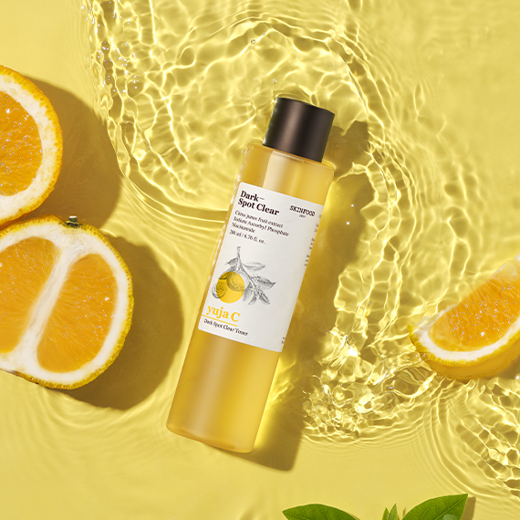 SKINFOOD Yuja C Dark Spot Clear Toner 200ml bottle surrounded by sliced yuja fruit and clear water ripples on a sunny yellow background.
