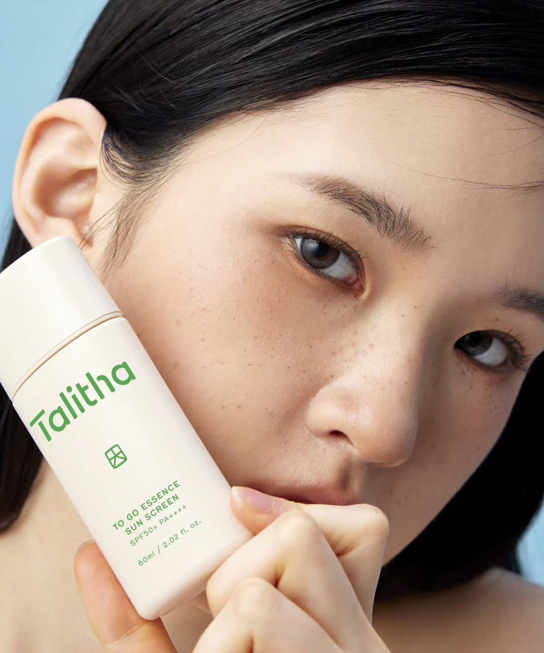Young woman holding a bottle of TALITHA KOUM To go Essence Sun Screen 60ml against her face.