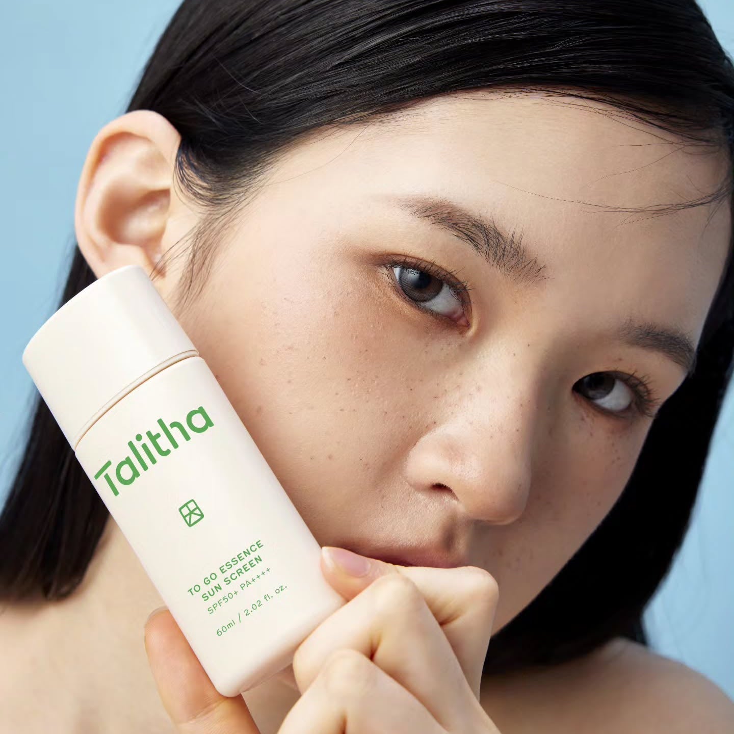 Young woman holding a bottle of TALITHA KOUM To go Essence Sun Screen 60ml against her face.