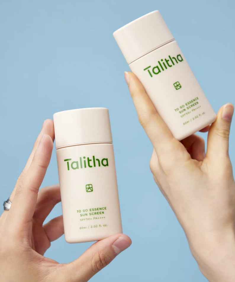 Two hands holding TALITHA KOUM To go Essence Sun Screen 60ml bottles against a blue background
