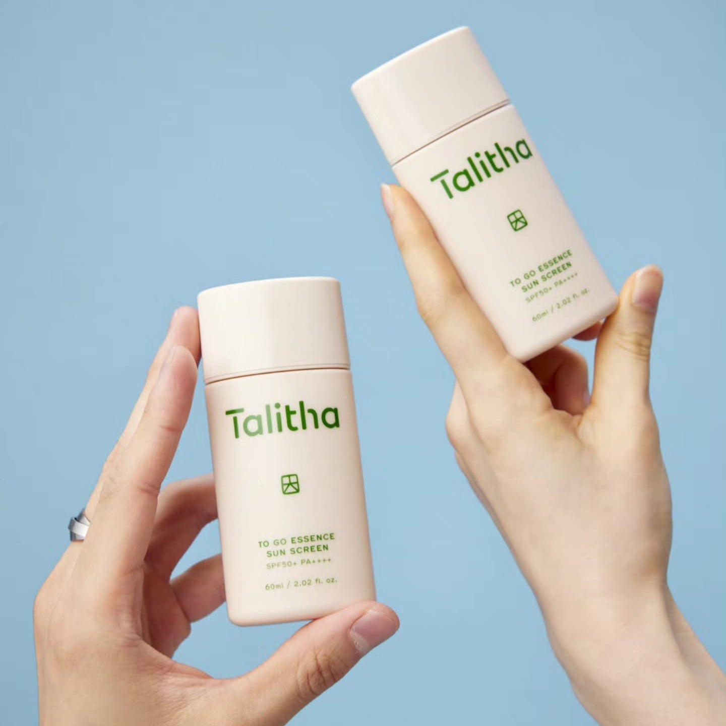 Two hands holding TALITHA KOUM To go Essence Sun Screen 60ml bottles against a blue background