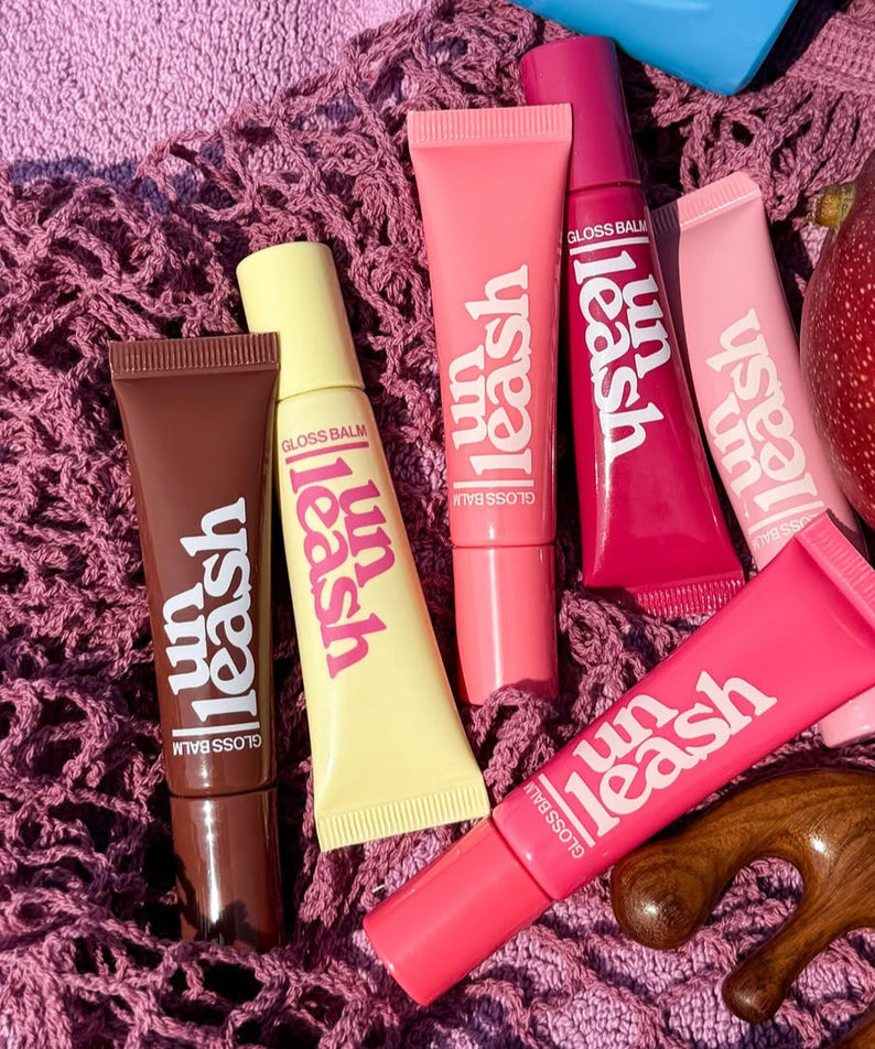 Collection of UNLEASHIA Sunset Dazzle Gloss Balm tubes in various shades displayed on a textured pink background.