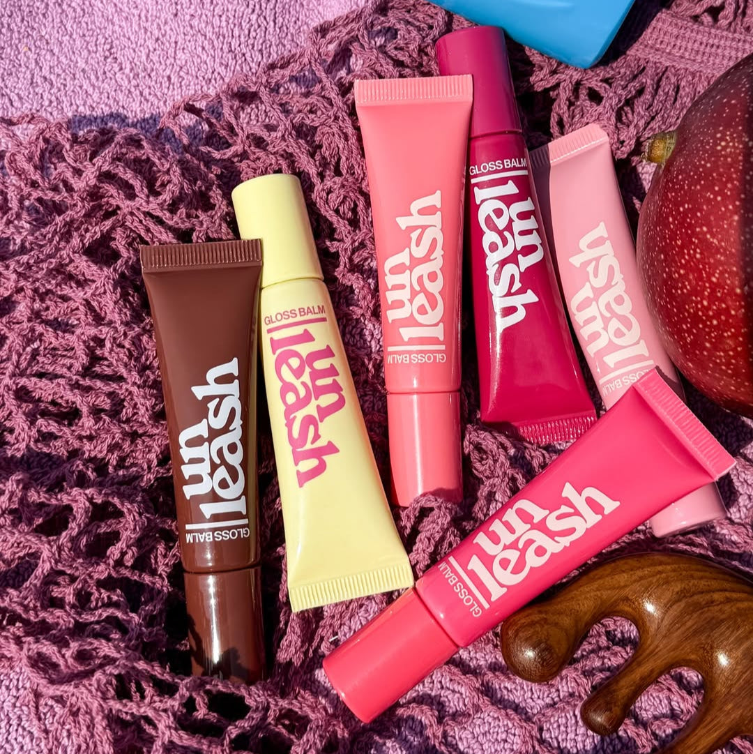 Collection of UNLEASHIA Sunset Dazzle Gloss Balm tubes in various shades displayed on a textured pink background.